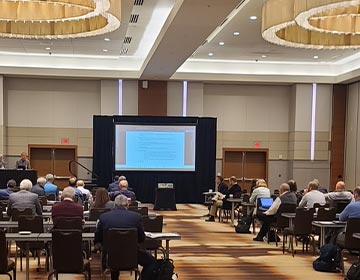 People seated in a large conference room attending a presentation displayed on a projector screen.